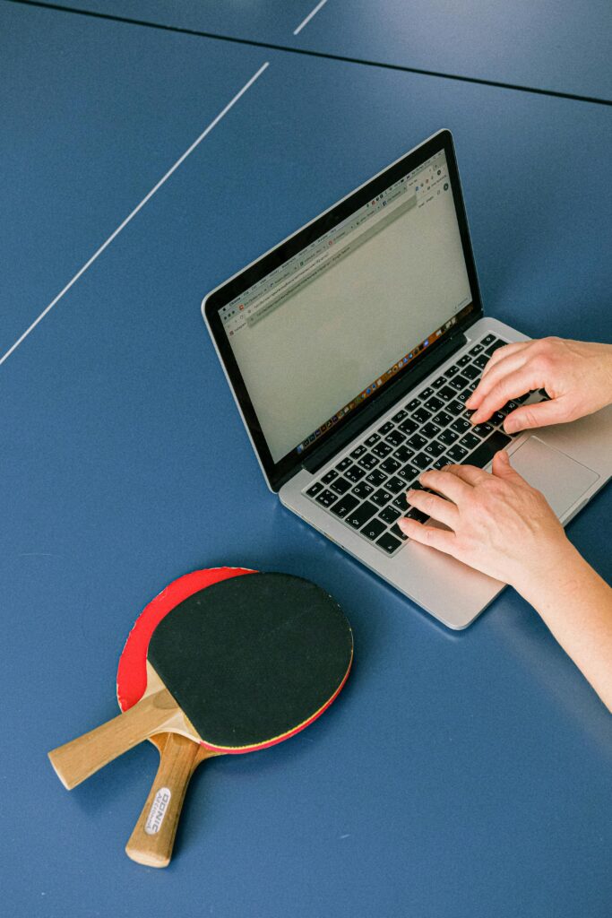 Hands typing on a laptop next to table tennis paddles on a blue table. Perfect blend of work and play.